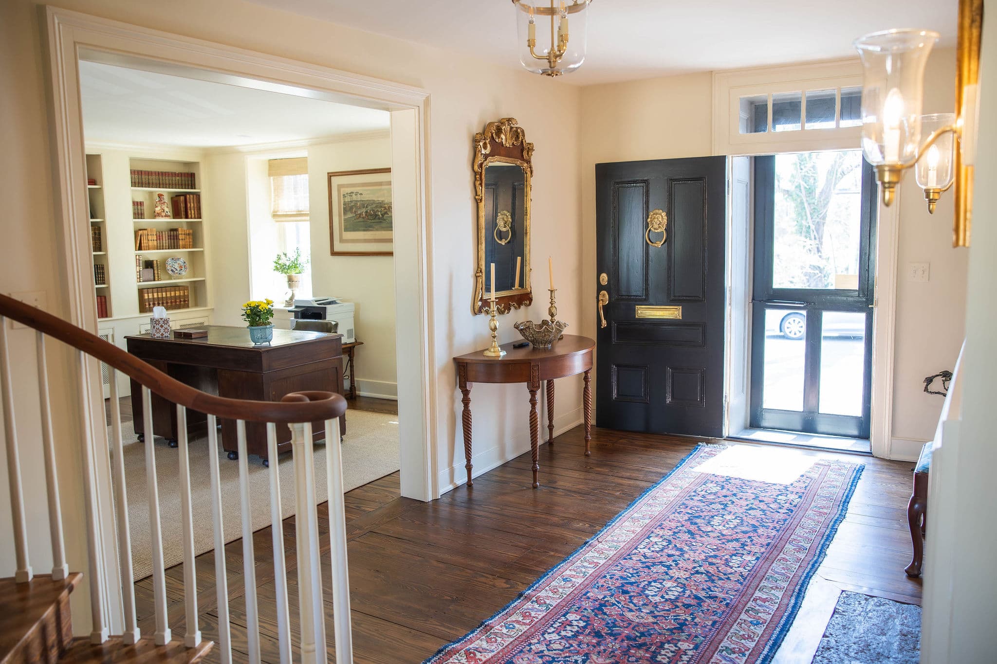 A bright entryway with a wooden staircase, decorative mirror, and an open front door leading to the outdoors.