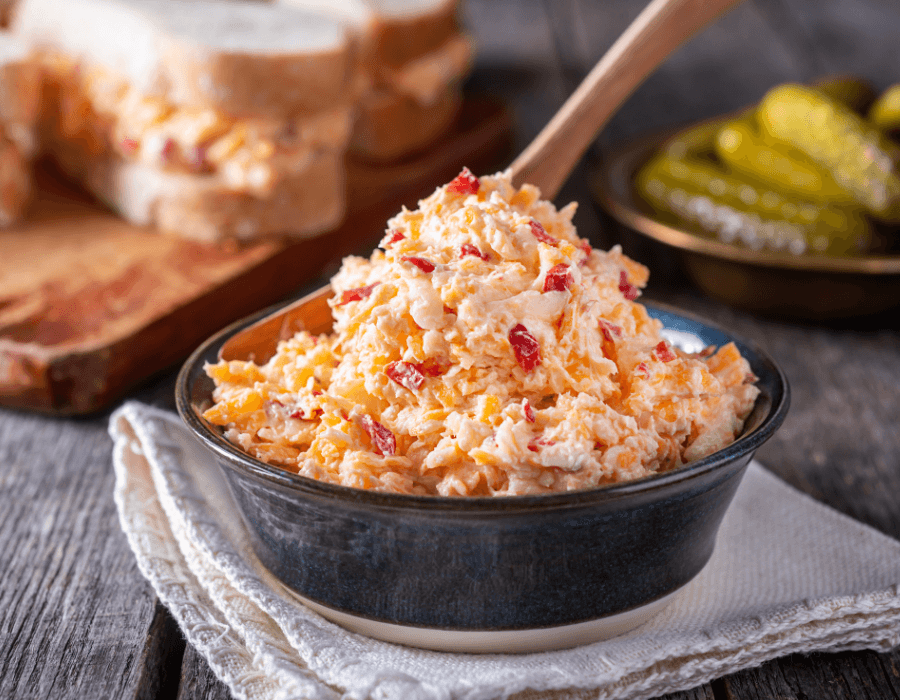 A bowl of pimento cheese spread with sandwiches and pickles in the background.