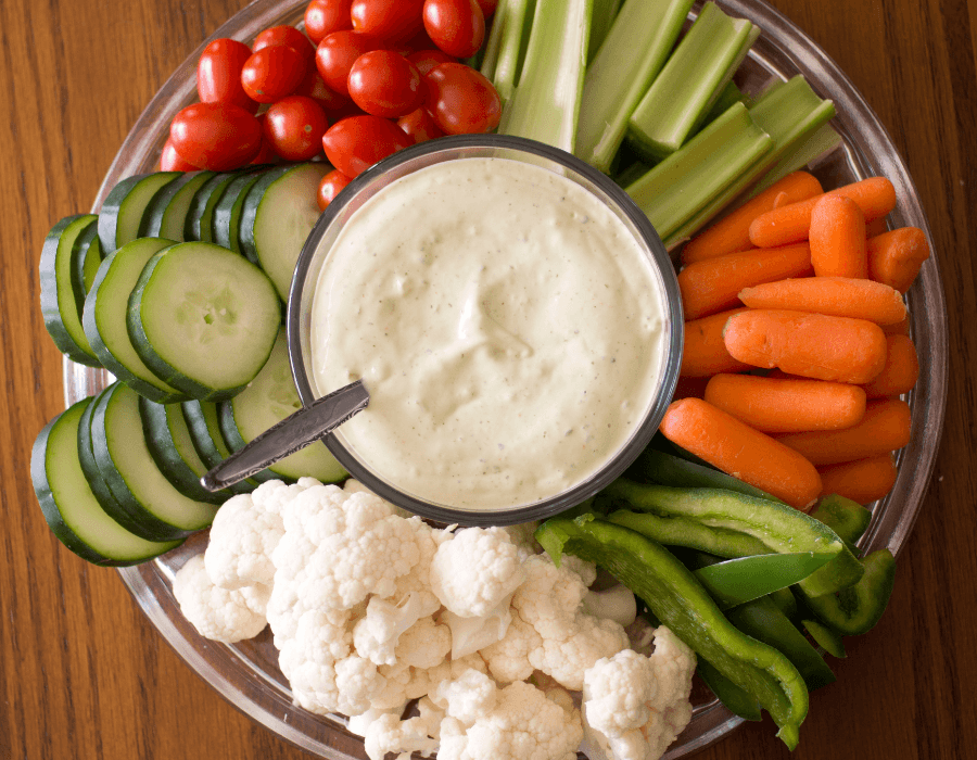 A platter of fresh vegetables including cherry tomatoes, celery, cucumber slices, carrots, cauliflower, and green bell pepper, served with a creamy dip in the center.