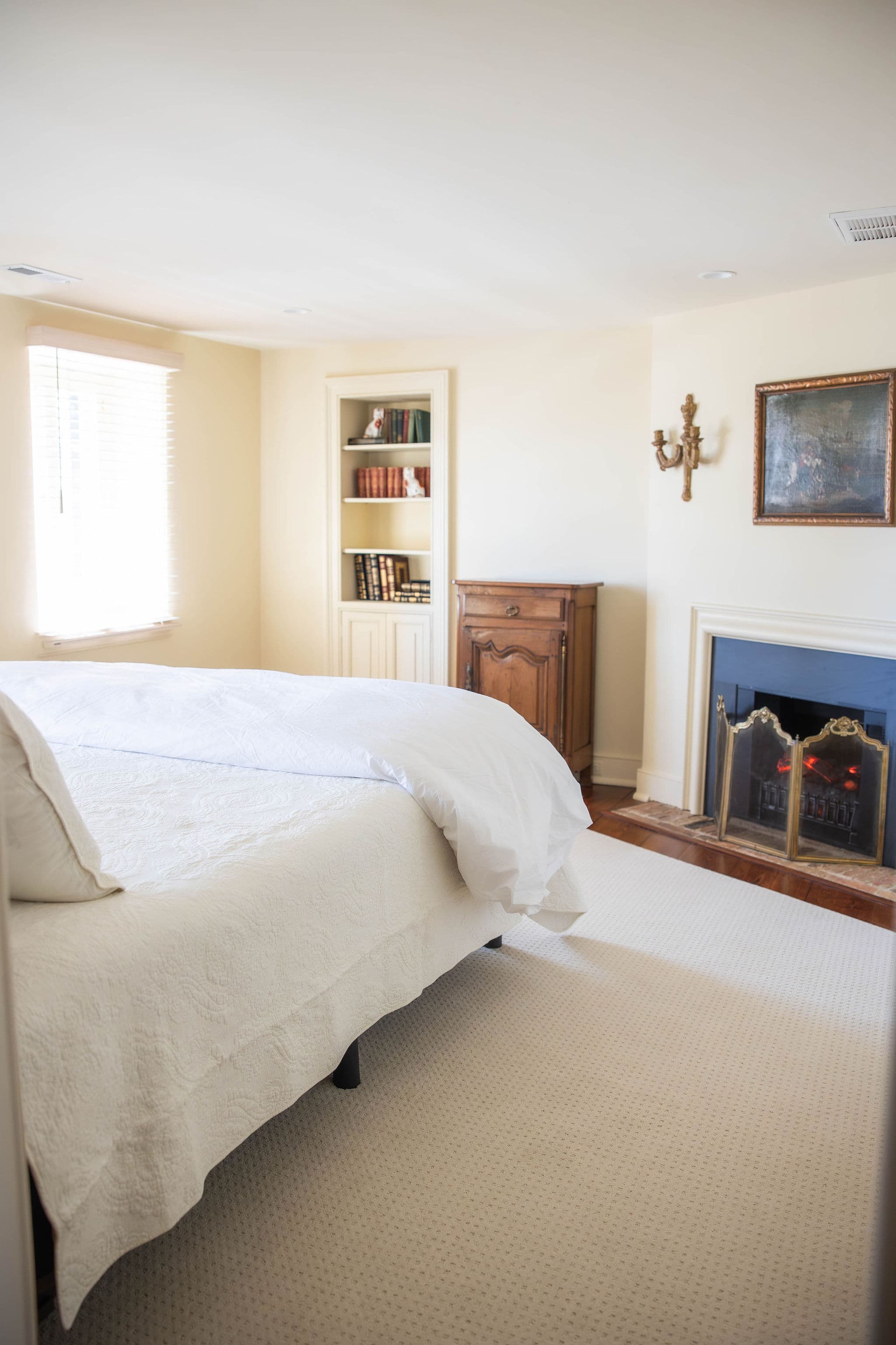 A cozy bedroom featuring a bed, a decorative fireplace, bookshelves, and natural light from a window.
