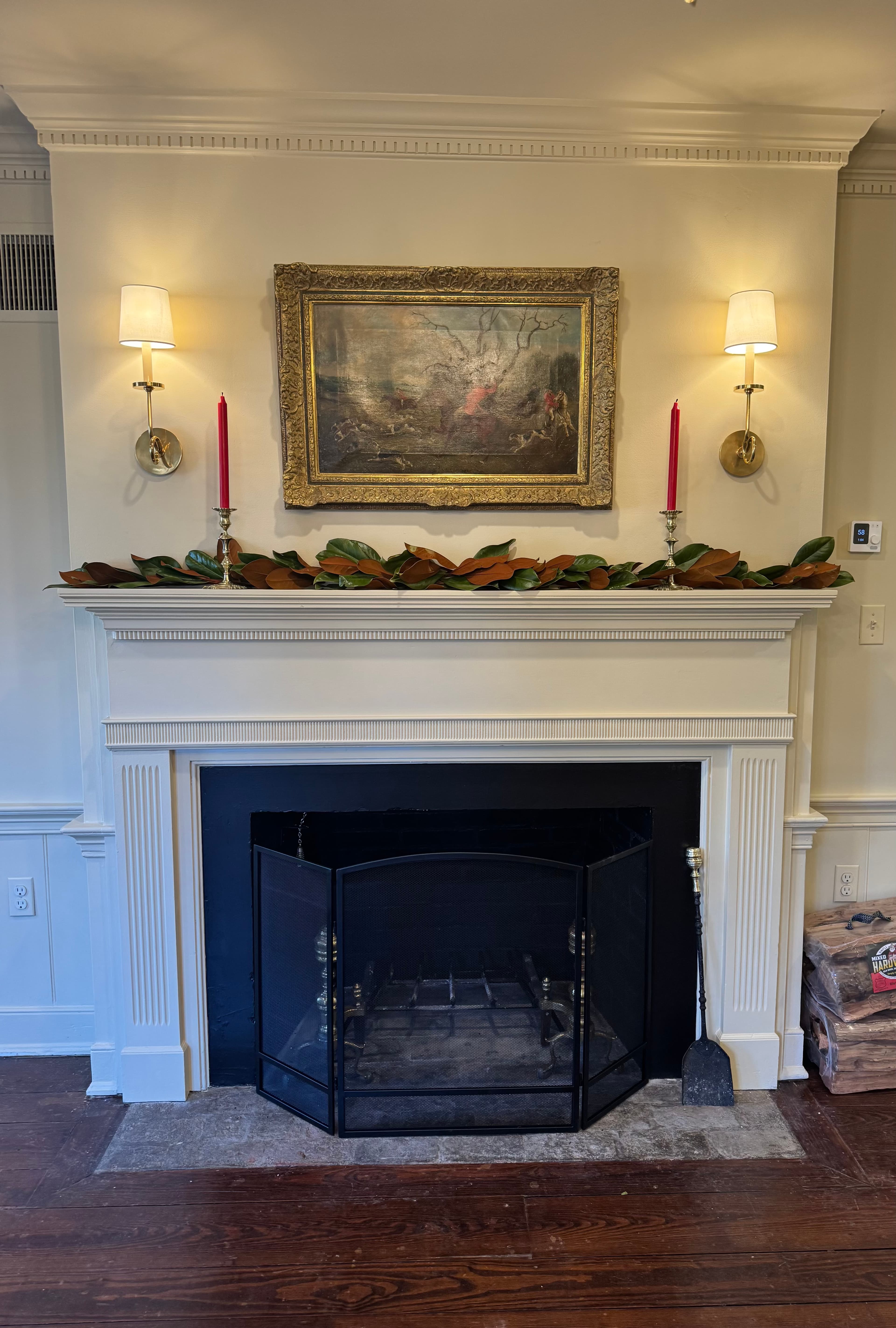 A decorated fireplace with a gold-framed painting, two wall sconces, red candles, and a garland.