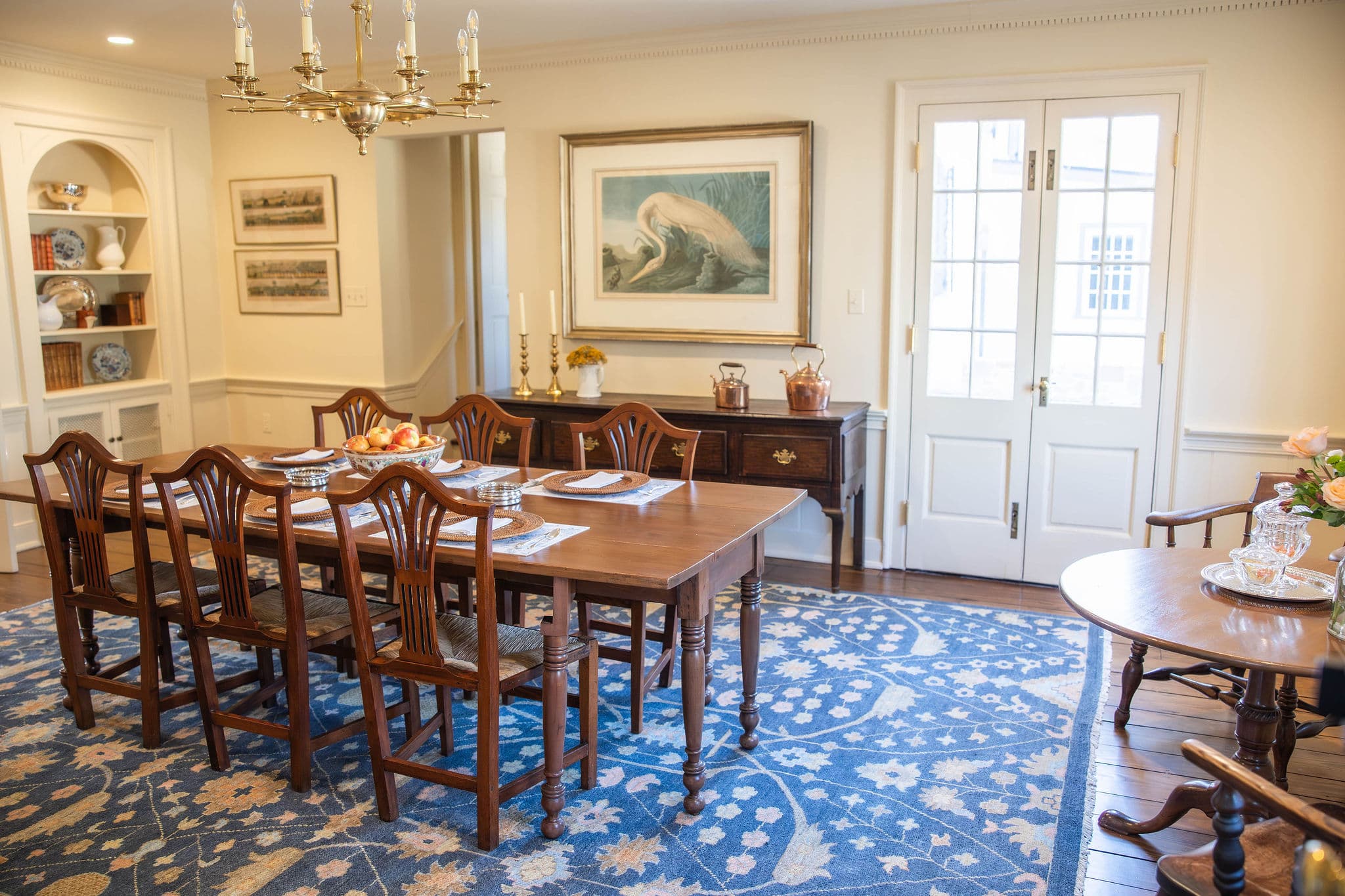 A formal dining room features a large wooden table, eight chairs, and an ornate chandelier, adorned with decorative artwork and a patterned rug.